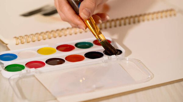 Partial view of woman holding paintbrush near watercolor on table 