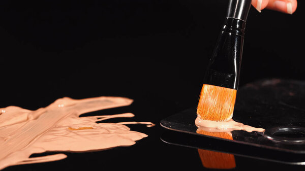 Close up view of woman holding cosmetic brush near face foundation on black background 