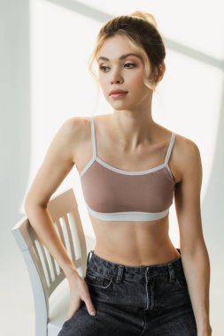 Spor ceketli zayıf bir kadın sandalyede poz veriyor ve gölgeli beyaz arka plana bakıyor.