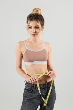 Spor elbiseli, sırıtan bir kadın. Bel ölçer. Gri üzerine izole bir şekilde başka yere bakar.
