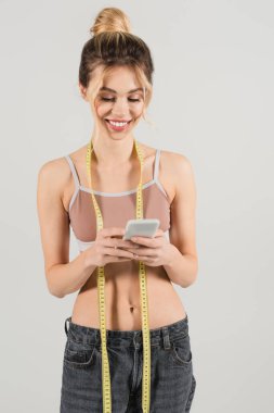 Gri üzerinde izole edilmiş cep telefonu kullanan ince vücutlu ve mezuralı neşeli bir kadın.