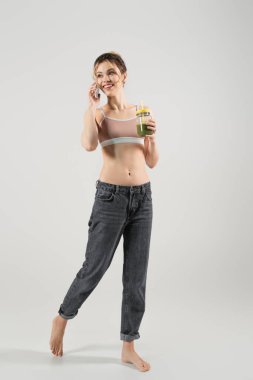 Smoothie dolu kavanozu olan çıplak ayaklı bir kadın gri arka planda akıllı telefondan konuşuyor.