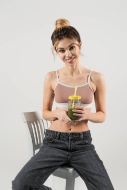 İnce vücutlu genç bir kadın sandalyede oturuyor. Gri üzerine izole edilmiş taze meyve suyu dolu bir kavanozla.