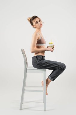 Spor bluzlu ve kot pantolonlu ince kadın, gri arka planda beyaz koltukta otururken smoothie tutuyor.