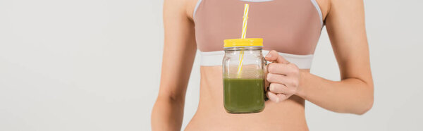 partial view of fit woman in sports top holding jar of fresh smoothie isolated on grey, banner