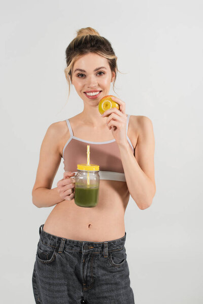 young woman with perfect face and body holding ripe apple and fresh smoothie isolated on grey