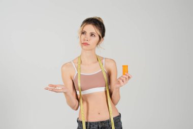 thoughtful woman with measuring tape holding vitamins and looking away isolated on grey clipart