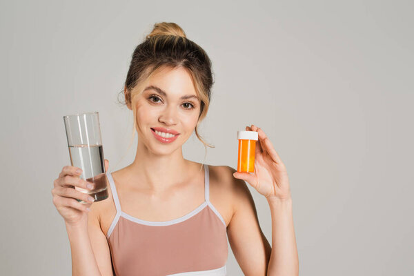 woman with perfect skin smiling at camera while holding glass of water and vitamins isolated on grey