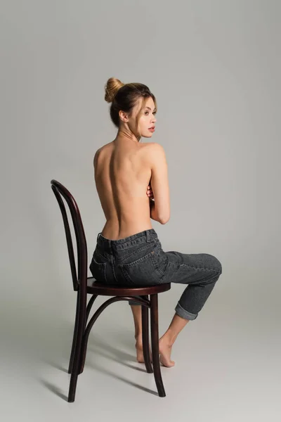 back view of half nude barefoot woman in jeans sitting on wooden chair and looking away on grey background