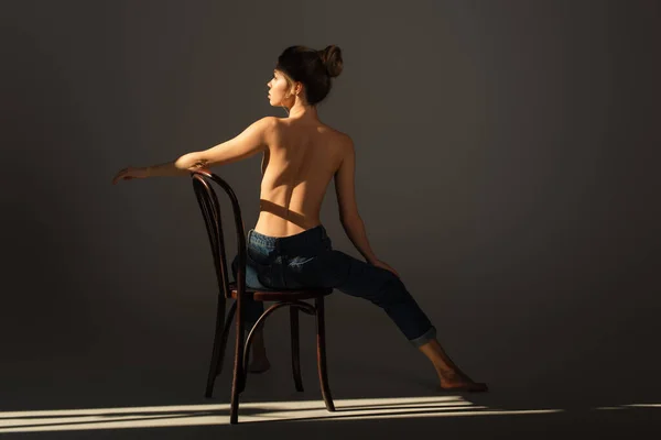back view of half nude barefoot woman in jeans posing on wooden chair on grey background with lighting