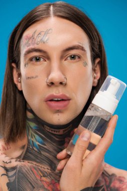 tattooed young man holding bottle with cleansing foam isolated on blue clipart