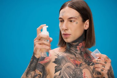 Genç ve dövmeli bir adam mavi şişeye temizleyici köpükle bakıyor.