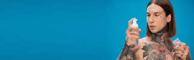 young and tattooed man looking at bottle with cleansing foam isolated on blue, banner clipart