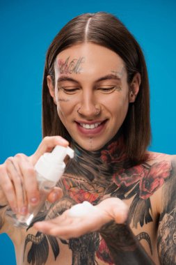 smiling and tattooed man holding bottle with cleansing foam isolated on blue clipart