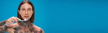 young and tattooed man holding toothbrush isolated on blue, banner clipart
