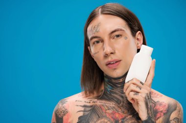 Piercingli ve dövmeli bir adam elinde kozmetik ürünlerle dolu bir şişe tutuyor. 