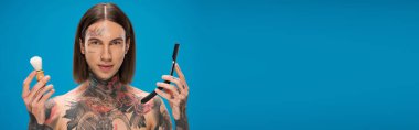 young and tattooed man holding shaving brush and mirror isolated on blue, banner clipart