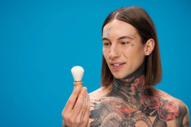 cheerful and tattooed man looking at shaving brush near face isolated on blue clipart