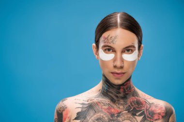 Üstsüz ve dövmeli genç bir adam. Göz bantlı. Mavi kameraya bakıyor.