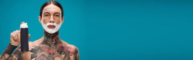 tattooed young man with shaving foam on face holding can isolated on blue, banner clipart