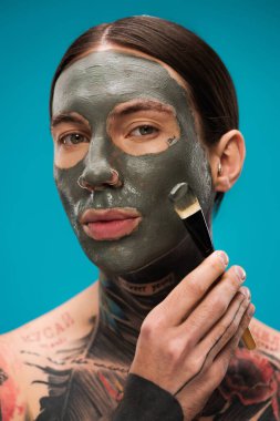 Üstsüz ve dövmeli bir adam. Mavi üzerine izole edilmiş kilden bir maske takıyor. 