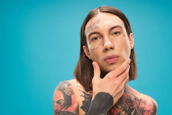 Jovem Perfurado Homem Com Tatuagens Tocando Rosto Liso Isolado Azul — Fotografia de Stock