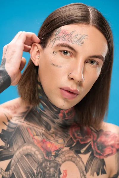 young and pierced man with tattoos looking at camera while adjusting hair isolated on blue