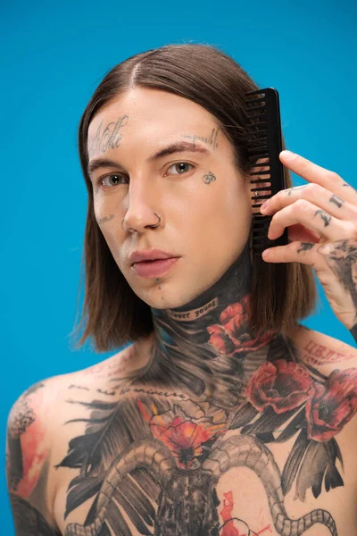young and pierced man with tattoos brushing hair with comb isolated on blue
