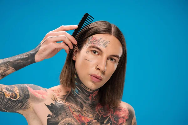 young and pierced man with tattoos brushing hair isolated on blue