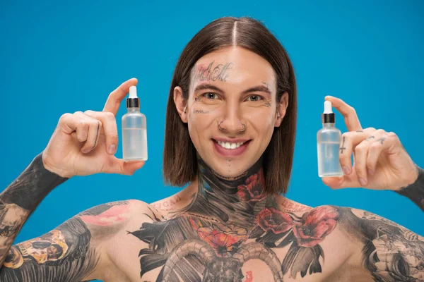 happy and tattooed man holding bottles with moisturizing serum isolated on blue
