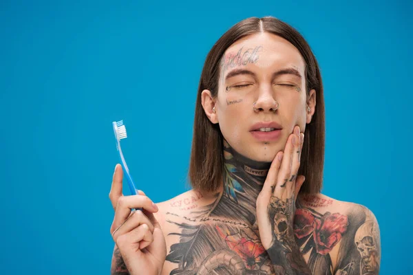 young and tattooed man with closed eyes holding toothbrush isolated on blue