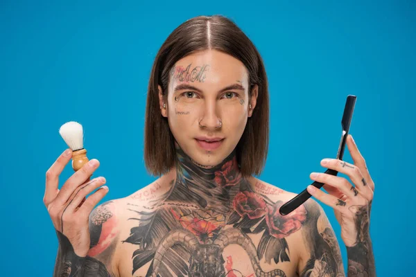 一个年轻的纹身男子拿着剃须刀和一面用蓝色隔离的镜子 — 图库照片