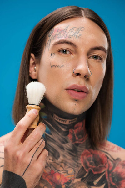 young and tattooed man holding shaving brush near face isolated on blue