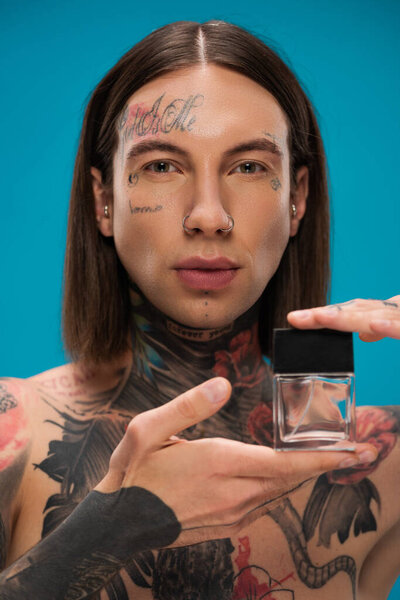 young and tattooed man holding bottle with perfume isolated on blue
