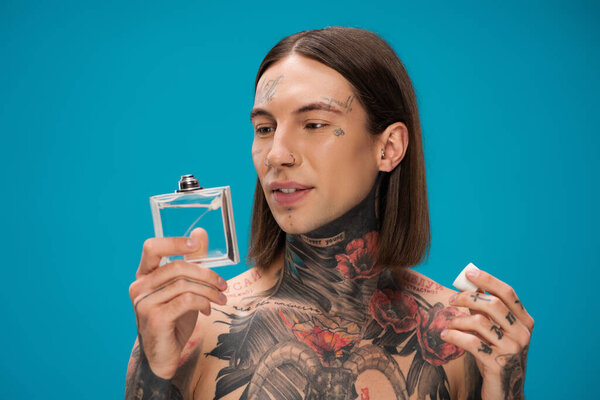 young and tattooed man looking at bottle with perfume isolated on blue