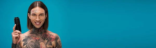 happy and tattooed young man holding antiperspirant isolated on blue, banner