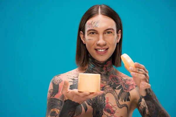 happy young man with tattoos holding container with body butter isolated on blue