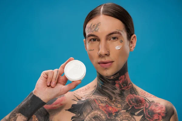 young man with tattoos and cream on cheek holding container isolated on blue