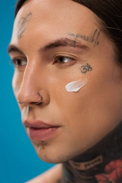 close up of pierced young man with face cream on cheek looking away isolated on blue