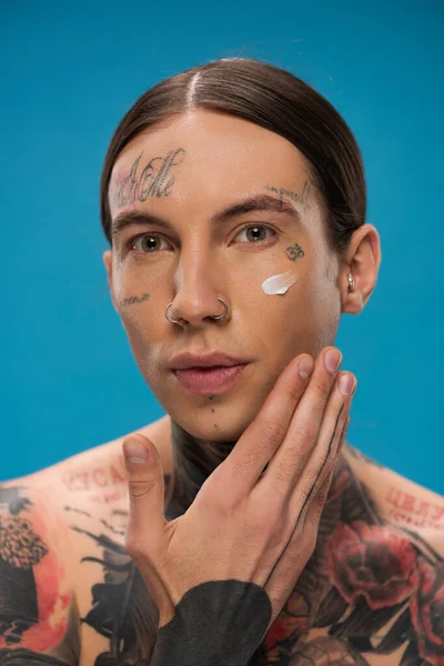 tattooed young man applying cream on face and looking at camera isolated on blue