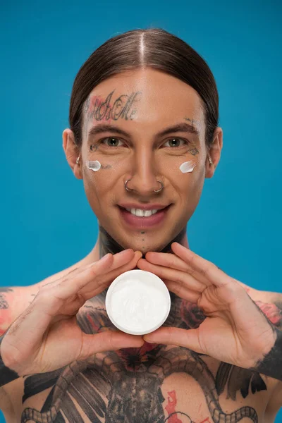 happy young man with tattoos and cream on cheeks holding container isolated on blue