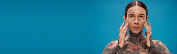 Татуированный Молодой Человек Наносит Крем Щеки Глядя Камеру Изолированную Голубом — стоковое фото