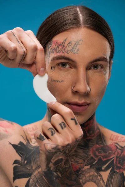 tattooed young man holding eye patch and looking at camera isolated on blue