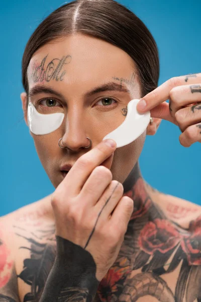 tattooed young man applying eye patches and looking at camera isolated on blue