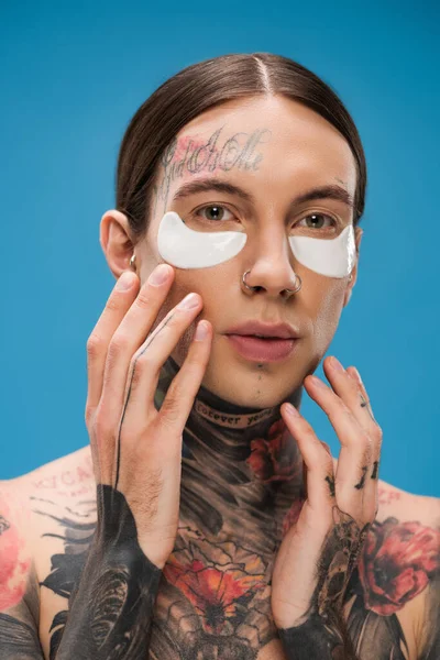 tattooed young man with eye patches looking at camera while touching face isolated on blue