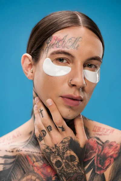 close up of tattooed young man with eye patches looking at camera while touching chin isolated on blue