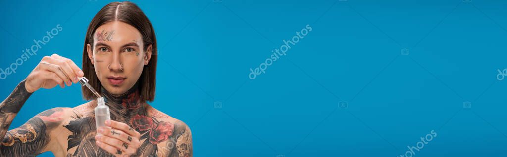 Young and tattooed man holding bottle with moisturizing serum isolated on blue, banner
