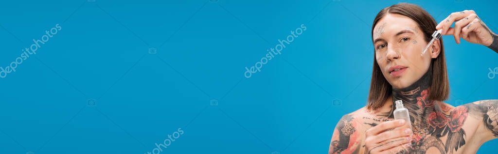 Young and tattooed man holding bottle while applying moisturizing serum isolated on blue, banner