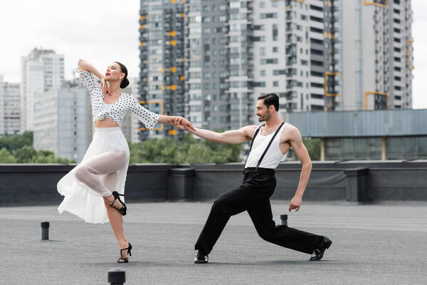 Улыбающийся мужчина держит партнера за руку во время танцев на крыше здания на открытом воздухе 