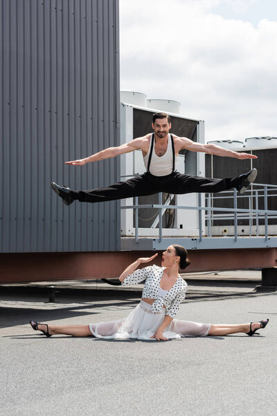 Smiling dancer doing split while jumping above shocked partner on roof of building outdoors 